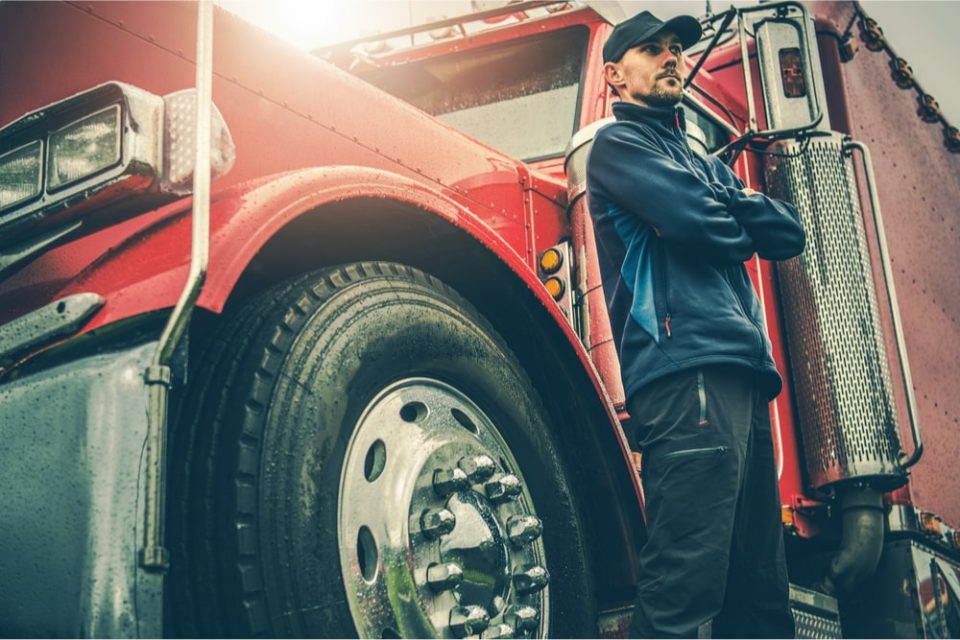 Truck driver in a blue uniform standing in front of a red semi-truck.