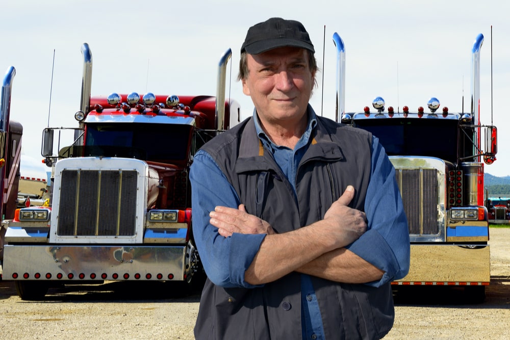 Middle aged man standing with his arms crossed, two semi-trucks in the background.
