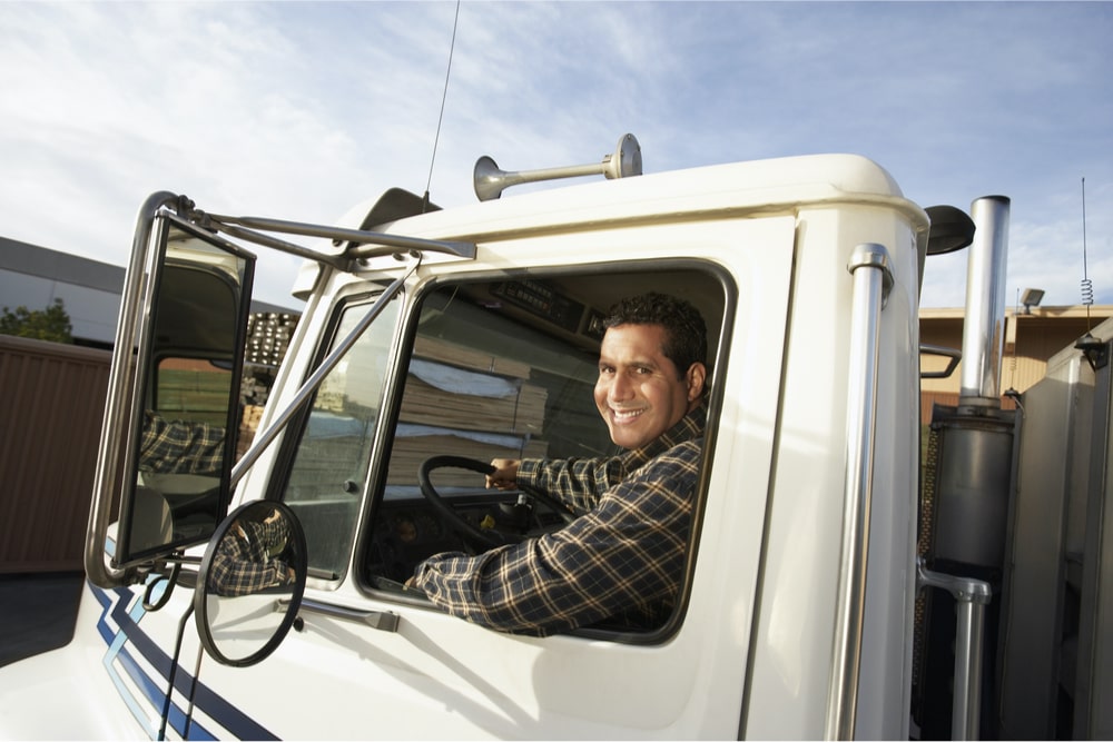 Man in dark plaid shirt looking out the driver's side window of a white semi-truck.