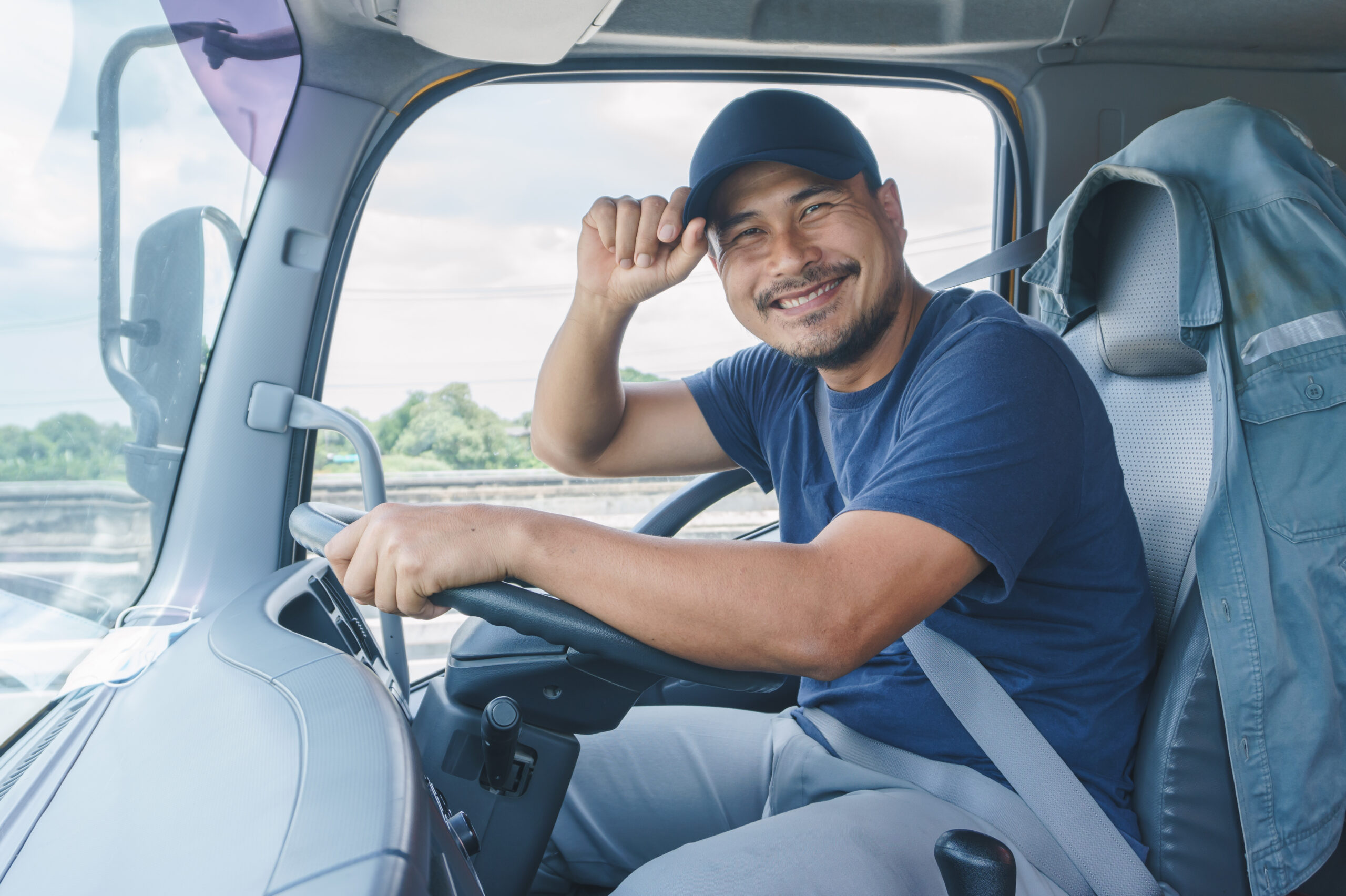 Male trucker in the cab of a truck facing the camera and tipping his baseball cap.
