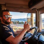Man wearing sunglasses and a beard giving a thumb's up in the cab of a truck.