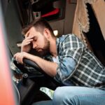 Bearded man wearing plain shirt in truck cab holding his hand to his head with his eyes closed.