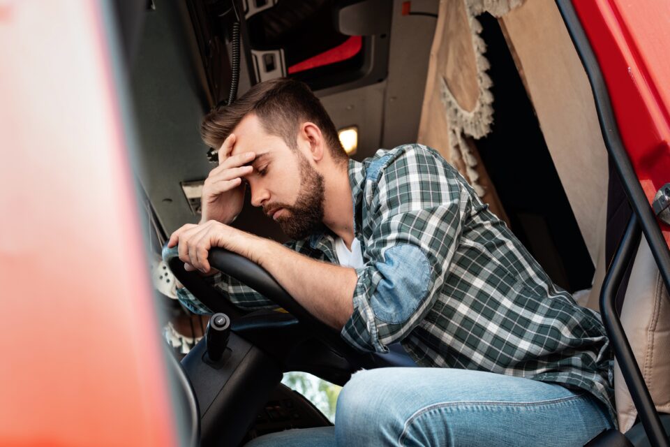 Bearded man wearing plain shirt in truck cab holding his hand to his head with his eyes closed.