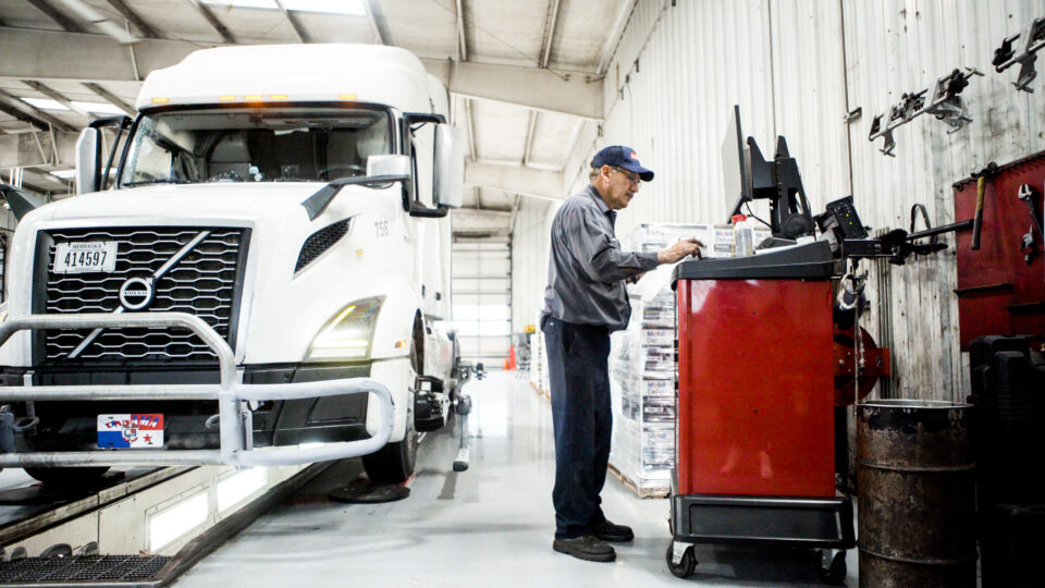 Mechanic using a computer workstation beside a white semi-truck in a service garage.
